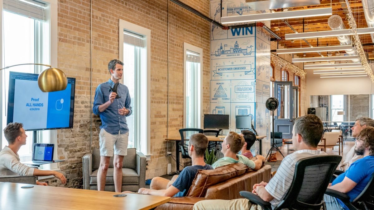 Team members in a modern office engaged in an all-hands meeting, with a presenter discussing a topic displayed on a screen