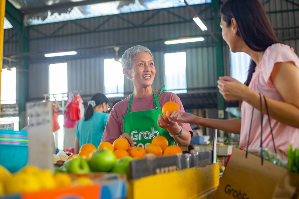 Smiling Grab delivery worker handing an orange to a customer at a vibrant market stall filled with fresh fruits