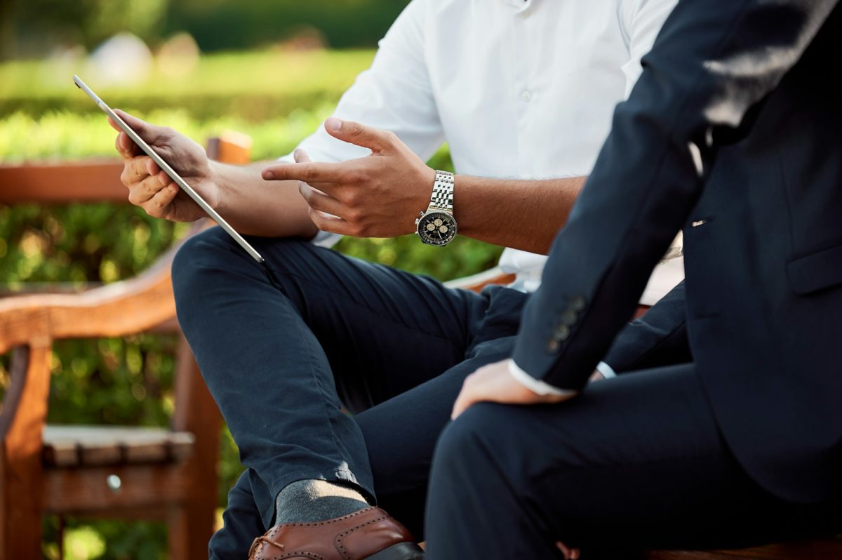 Business professionals discussing ideas on a tablet while sitting on a bench outdoors in a park-like setting