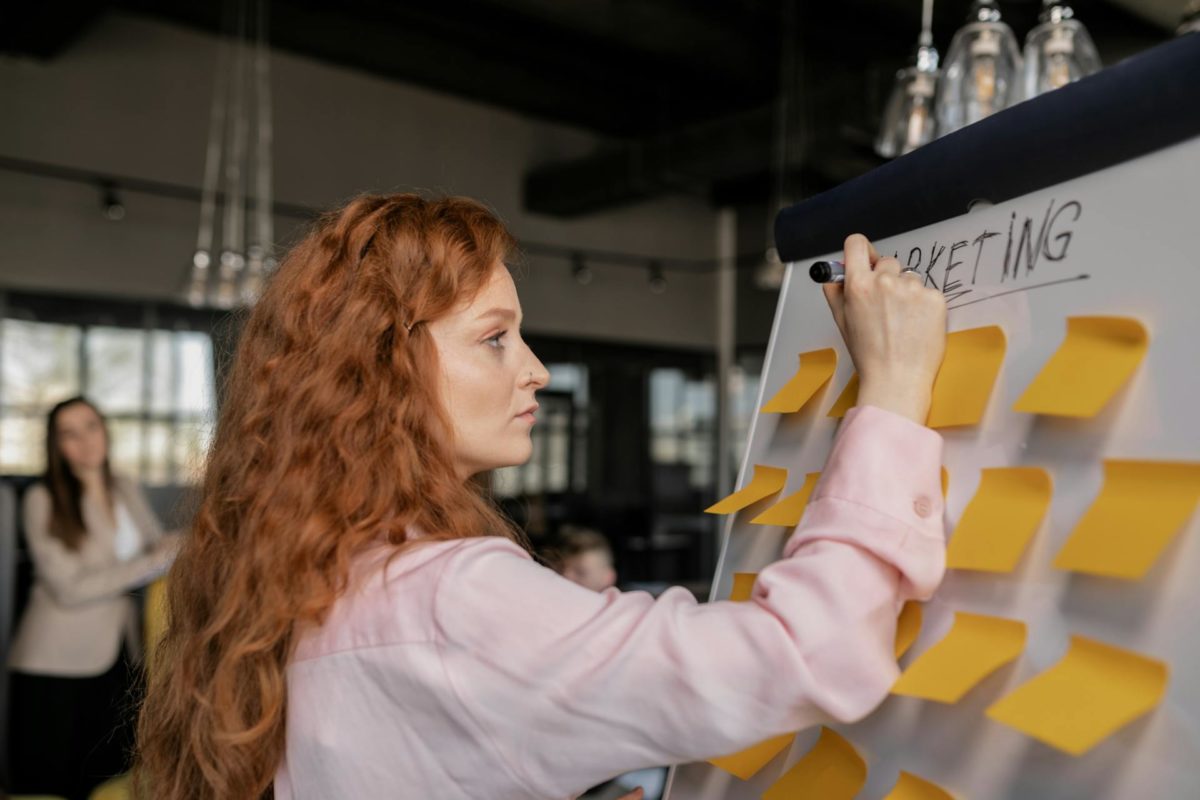 Businesswoman with long red hair writing on a whiteboard during a marketing strategy session, with sticky notes and colleagues in the background